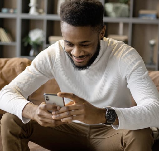 A man in his living room is smiling while texting on his smartphone.