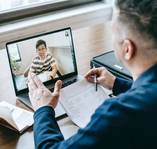 A man on his desktop is writing on different papers during a meeting with an other person on his laptop.