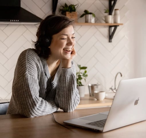 A woman in her kitchen is smiling during a video call on her laptop.